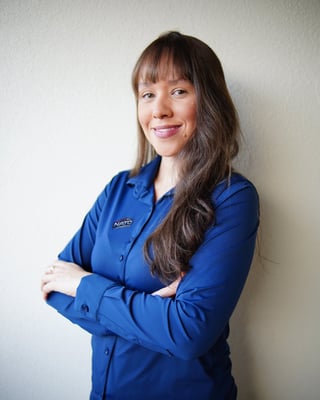 Professional headshot of a woman with long dark hair and bangs, wearing a blue work shirt, smiling at camera with arms crossed