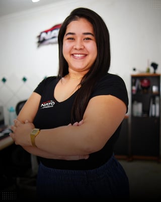 Smiling woman with long black hair wearing a black NATO branded t-shirt, standing with arms crossed in an indoor office setting