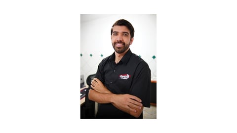 Man with dark hair and beard wearing black polo shirt with logo, posing with arms crossed against white background